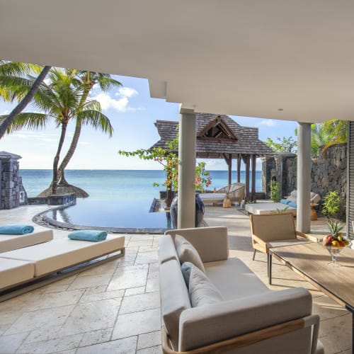 Luxurious open-air living area with modern wooden furniture and light blue cushions overlooking an infinity pool and tropical garden with palm trees by the sea.