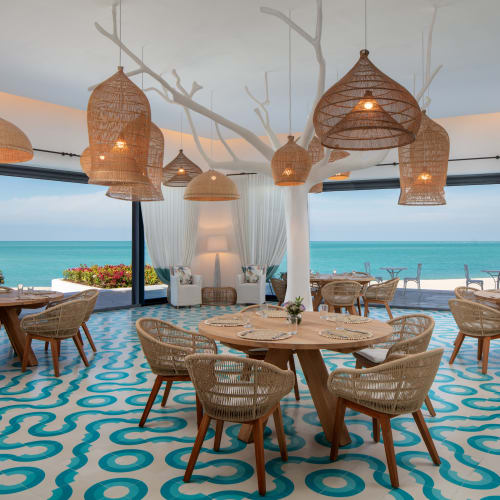 Elegant interieur eines Restaurants mit Blick auf den Ozean durch große Glastüren, Holztische und moderne geflochtene Lichtinstallationen.