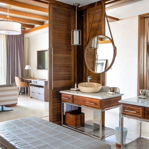 Luxuriöses Hotelzimmer mit Holzakzenten, modernen Waschbecken mit runden Spiegeln und einem geräumigen Wohnbereich mit großen Fenstern und bequemen Sitzgelegenheiten.