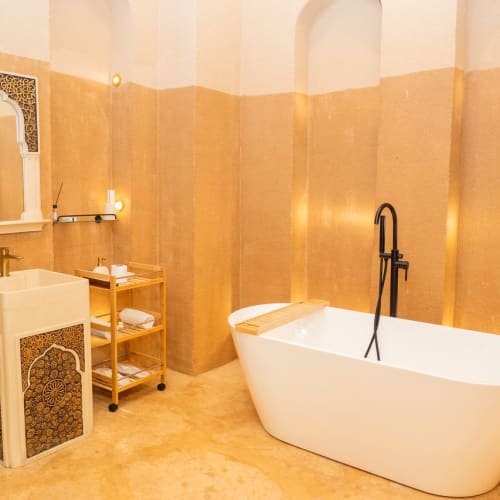 Ein Badezimmer mit einer freistehenden weißen Badewanne, einem weißen Waschbecken mit aufwendigen dunklen Musterpanelen, einem verzierten Spiegel und einem kleinen hölzernen Regal mit Handtüchern vor einer warmen beigefarbenen Wand.