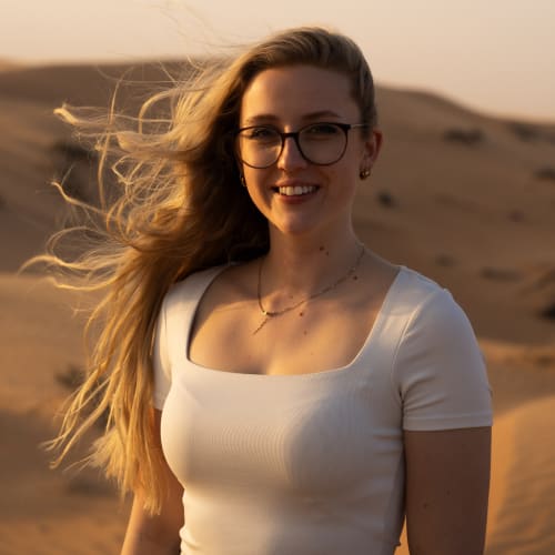 Young woman with long blonde hair and glasses standing in a desert with sand dunes at sunset, wearing a white top and smiling.
