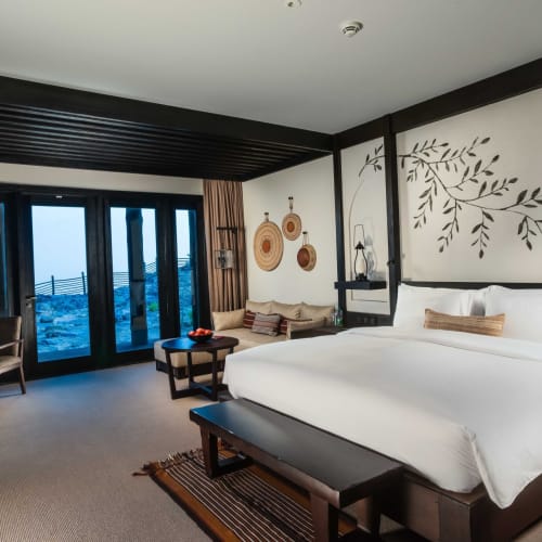 A luxurious hotel room with a large bed, a sitting area with a sofa, and large glass doors leading to a balcony with a mountain view.