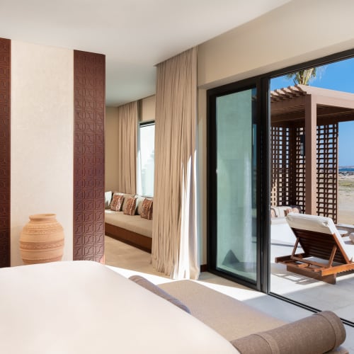 Luxurious hotel bedroom with a large bed and sliding glass doors opening to a terrace with a pool and beach view.