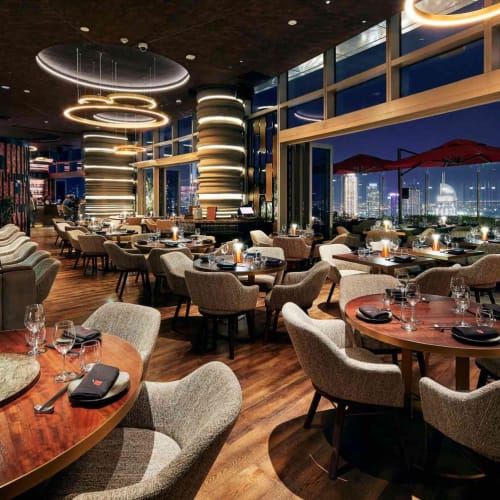 Elegantes Restaurant mit modernster Beleuchtung, runden Tischen, bestückt mit Besteck, und großen Fenstern, die eine Stadtansicht bei Nacht zeigen.