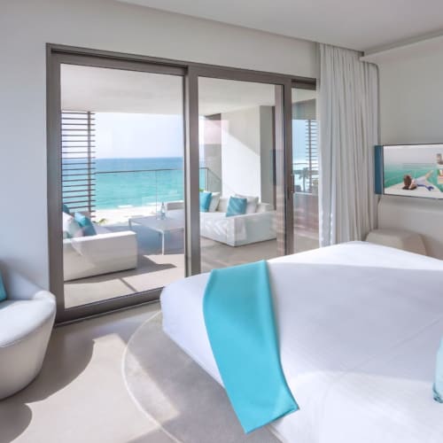A bright hotel room with a large bed, turquoise accents, and a view of the ocean through sliding glass doors leading to a balcony.