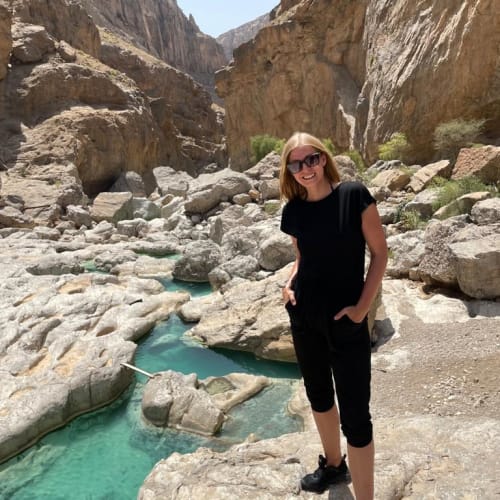 Eine Frau mit schwarzer Kleidung und Sonnenbrille steht auf felsigem Gelände neben einem klaren blauen Bach in einer engen Bergschlucht unter klarem Himmel.