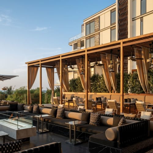 A stylish outdoor lounge area with modern furniture and a canopy, overlooking the cityscape at sunset.