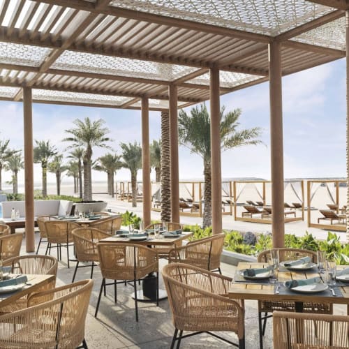 An open-air restaurant with wooden furniture and tables set for dining, overlooking a beach with palm trees and lounge chairs.