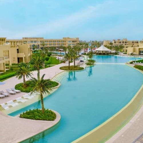 Luxurious resort pool with palm trees and modern buildings in the background on a sunny day.