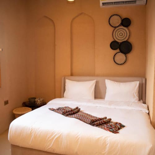 Ein gemütliches Schlafzimmer mit einem großen Bett, das mit weißen Bettlaken und einem dekorativen gemusterten Läufer bedeckt ist, vor einer beigen Wand mit ornamentalen Aussparungen und Wanddekorationen, neben einem geöffneten Holzfenster.