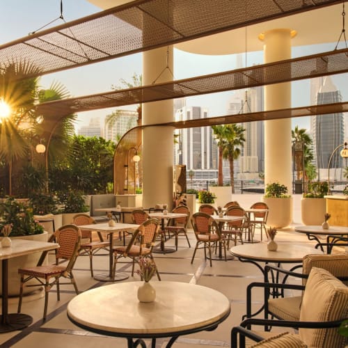 Elegante Außenterrasse eines Restaurants mit runden und rechteckigen Tischen, Korb- und gepolsterte Stühle sowie üppiges Grün mit Palmen, mit Blick auf die Skyline.