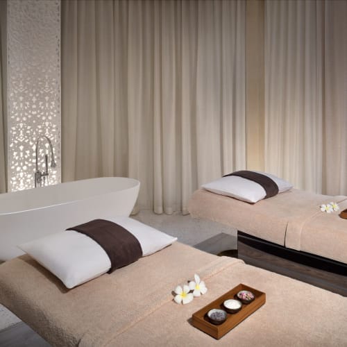 Zwei Massageliegen mit Handtüchern und Blumenbedaken in einer Wellness-ähnlichen Umgebung, mit Badewanne und Vorhängen im Hintergrund.