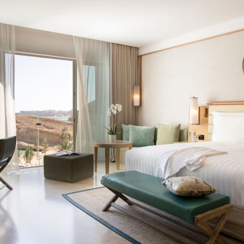 A luxurious hotel room with a large bed, green and white decor, and a balcony view of the ocean and rocky landscape.