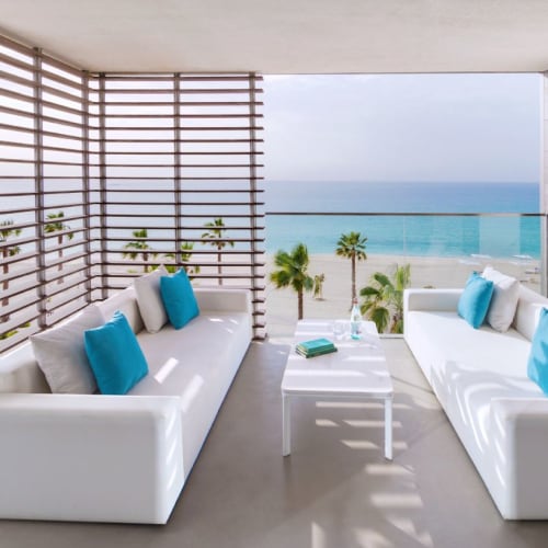 A modern balcony with white sofas and blue cushions overlooking a beach with palm trees and the ocean.