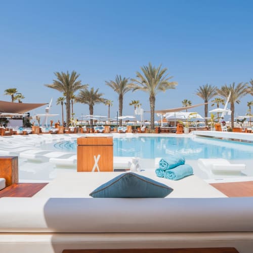 Luxuriöse Poolseite mit Nikki Beach Resort und Spa Dubai, Palmen und Sonnenliegen unter Sonnenschirmen.