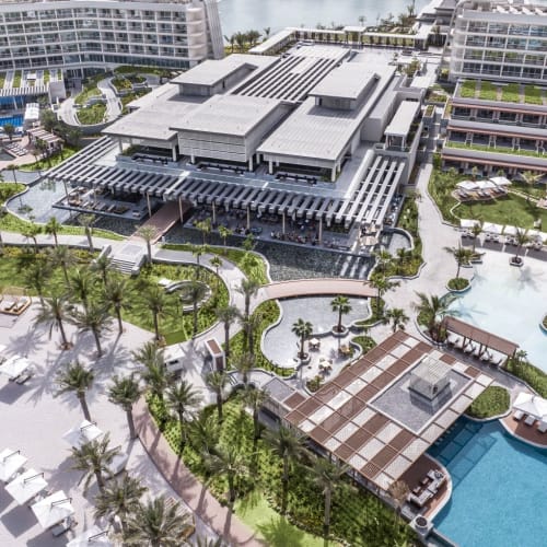 Luftaufnahme eines Luxusresorts mit großem Swimmingpool, Palmen, mehreren Gebäuden und einem Strandbereich.