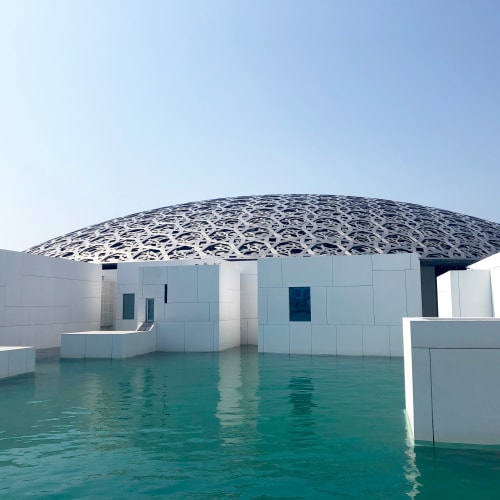 The Louvre Abu Dhabi's distinctive dome structure over white buildings with reflections in water.
