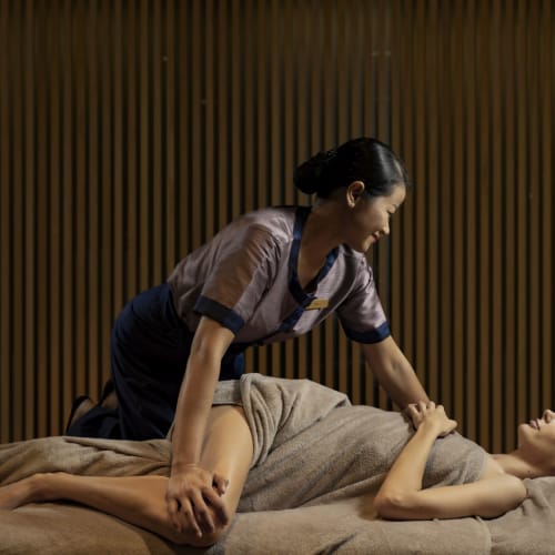 Eine Massagetherapeutin gibt einer liegenden Frau in einem Wellnesszentrum eine Massage, vor einer Holzverzierung im Hintergrund.
