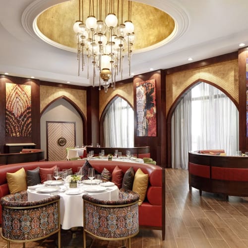 Elegant dining area in a luxurious restaurant with a round table set for a meal, ornate chairs, arched windows, and vibrant artwork on the walls.