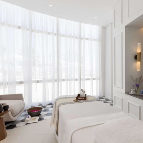 A spacious hotel room with large floor-to-ceiling windows, white curtains, and modern minimalistic decor including a bed, a sitting area with a sofa, and a sink area with a mirror.
