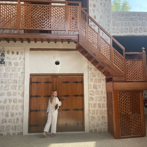 A woman in white pants and a white top stands near large wooden doors in a stone building with a balcony and staircase.