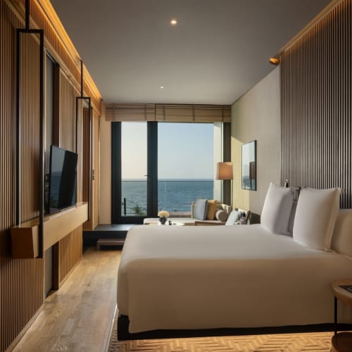A modern hotel room with wooden wall panels, a large bed, and a sea view through floor-to-ceiling windows.