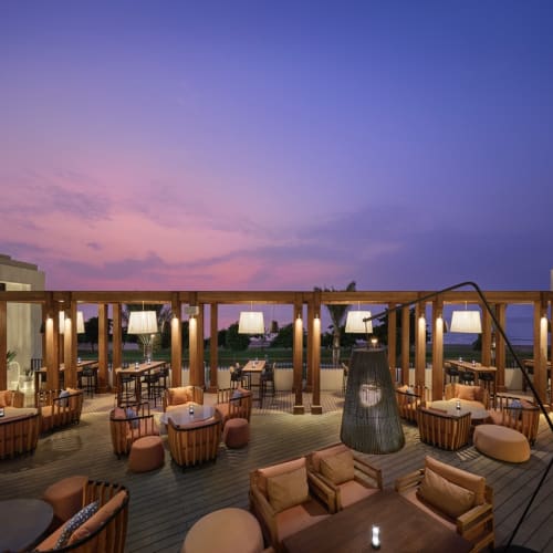 Eine stilvolle Outdoor-Lounge mit bequemen Sitzgelegenheiten um einen großen Baum, beleuchtet durch hängende Pendelleuchten und Korblampen.