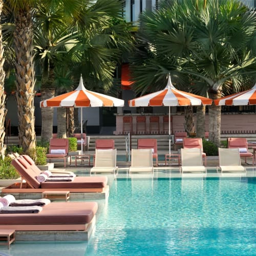 Sonnenliegen und Sonnenschirme rund um einen Swimmingpool in einem luxuriösen Resort, umgeben von hohen Palmen und üppigem Grün.