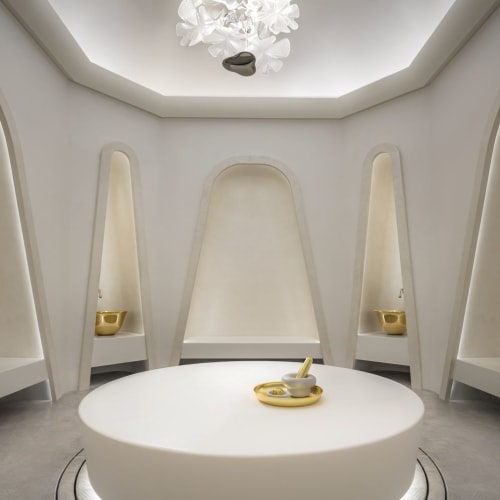 Ein futuristischer, minimalistischer Raum mit geschwungenen weißen Wänden, einem zentralen weißen Tisch mit einer goldenen Schüssel und fünf schmalen Wandnischen mit ähnlichen goldenen Schüsseln.