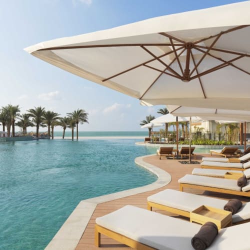Luxuriöse Poolseite mit Sonnenliegen und großen Sonnenschirmen, Blick auf das Meer, im Hintergrund Palmen.