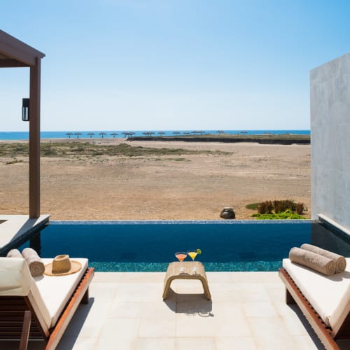 Entspannter Terrassentisch mit zwei Liegestühlen und kleinem Tisch mit Blick auf Strand mit ruhigem Meer und blauem Himmel.