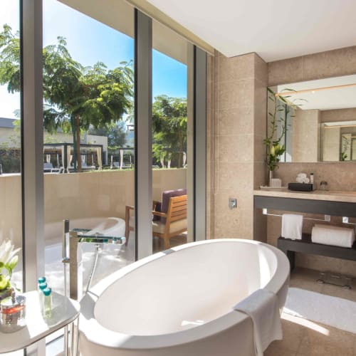 Luxuriöses Hotelzimmer mit freistehender Badewanne, großem Spiegel und Blick nach draußen durch GlasschiebeTüren.