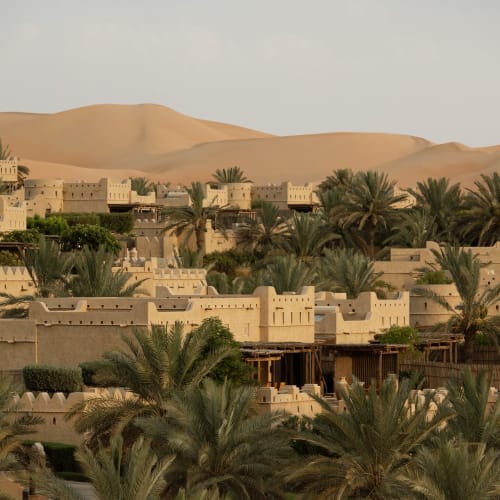Ein Wüstenresort mit traditioneller Architektur, umgeben von Palmen, vor sandigen Dünen in einer Wüstenlandschaft.