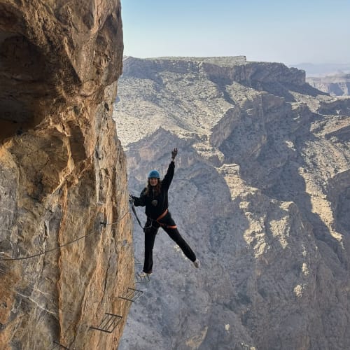Person klettert an einer steilen felsigen Klippe mit Eisenklammern und Seilen einer Via Ferrata in einer bergigen Wüstenlandschaft unter klarem blauem Himmel.
