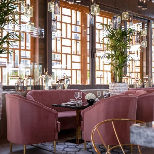 Elegantes Restaurantinnenraum mit rosa gepolsterten Stühlen, großen Topfpflanzen und großen Fenstern mit Holzrahmen, die natürliches Licht hereinlassen.