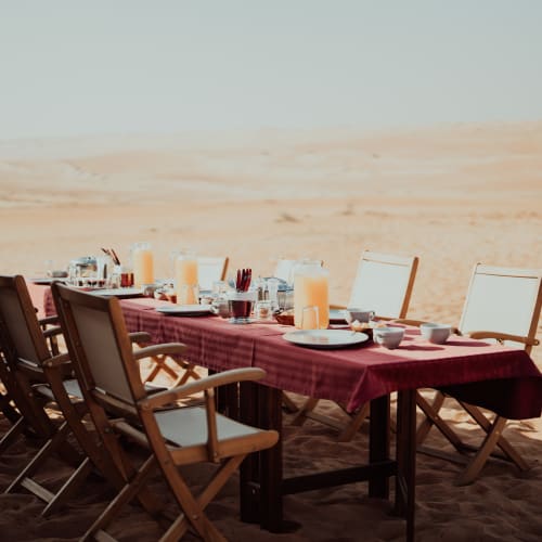 Ein langer Holztisch mit einem roten Tischtuch, gedeckt zum Frühstück mit Saftkrügen, Tassen und Tellern in einer sandigen Wüstenlandschaft bei hellem Tageslicht.