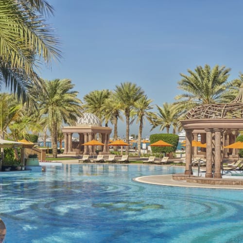 Luxuriöse Poolanlage mit Palmen, schattigen Kabinen und orangefarbenen Sonnenschirmen unter klarem blauen Himmel.