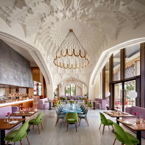 Elegantes Restaurant-Innenraum mit hoher, verzierter weißer Decke, bunten Stühlen um Tische und großen Fenstern, die natürliches Licht hereinlassen.