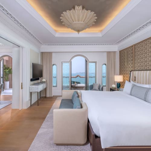 Elegant hotel rooms with large windows, sea view, a large double bed, and living room furniture. The design is very elegant in white and gold tones.
