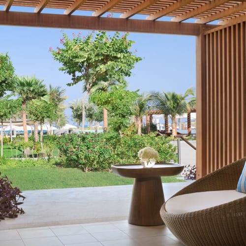 A cozy indoor seating area with a view of a lush garden and palm trees outside, under a wooden pergola.