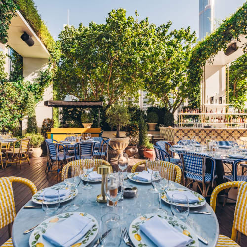 An outdoor dining area on a wooden deck, decorated with colorful chairs, table settings, and lush greenery.