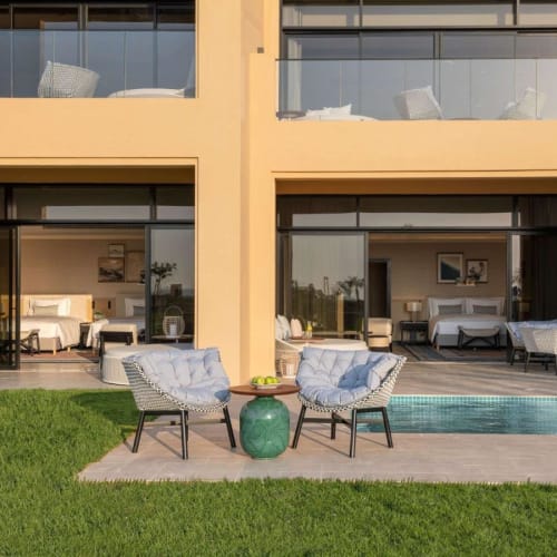 Two outdoor chairs with cushions and a small table sit on a patio in front of a modern building with glass doors.