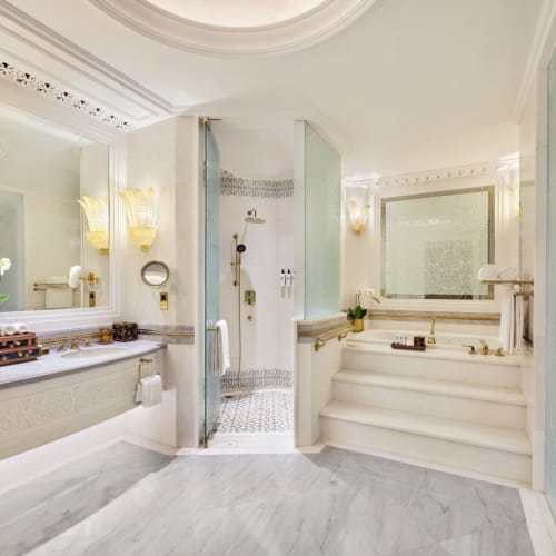 Luxuriöses Hotelbad mit weißen Marmor-Böden, eleganten Spiegeln und Goldakzenten, mit einer Dusche und einer Treppe, die zu einer Wanne führt.