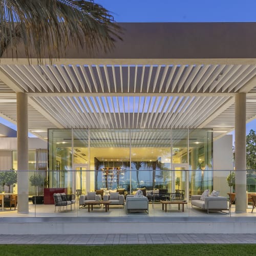 Modern beachfront resort with a glass-walled lounge and outdoor seating area under a large overhanging roof, overlooking greenery and water.