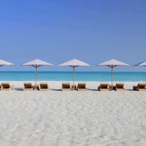 Seven beach chairs with umbrellas lined up on a sandy beach facing the calm sea and clear blue sky.