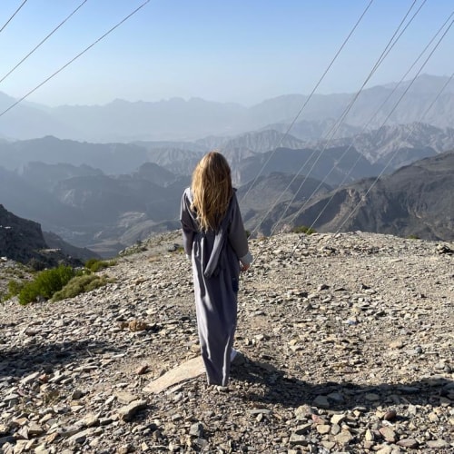 Eine Frau steht auf einem felsigen Berggelände mit bergigem Panorama im Hintergrund unter klarem Himmel.