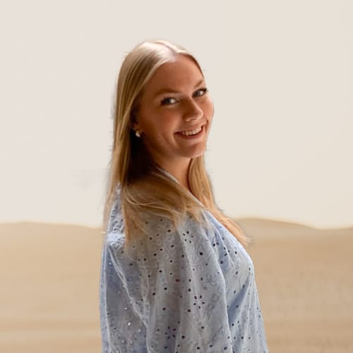 A smiling young woman with blonde hair standing indoors with a neutral background.