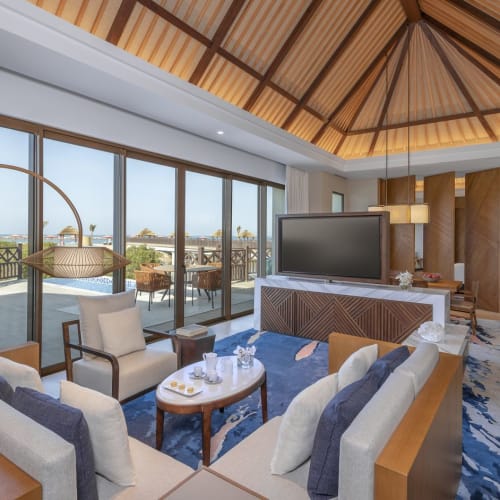 Luxuriöses Hotelwohnzimmer mit großen Schiebetüren, die zu einem Balkon mit Meerblick führen, mit modernen Möbeln und einer hohen Holzdecke.