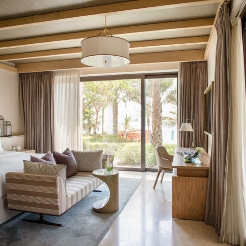 Luxushotelzimmer mit großem Bett, einem gestreiften Sofa und Schreibtisch, mit Panoramafenstern und Blick auf den Garten.