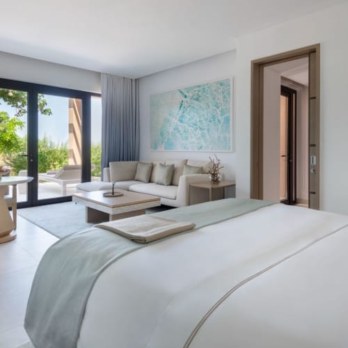 Modernes Hotelzimmer mit großem Bett, Sitzecke mit Sofa und Stühlen sowie Balkon mit Blick auf die Natur.